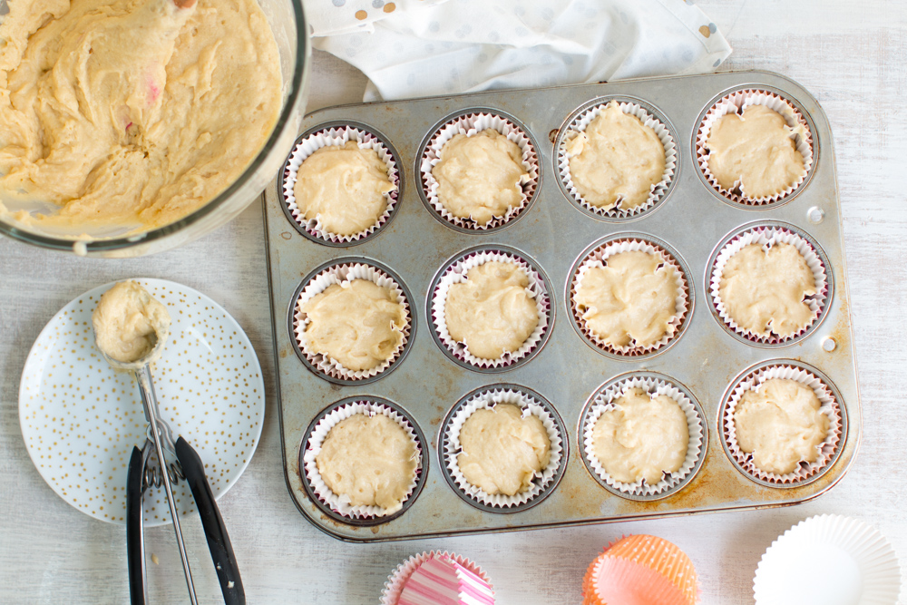 a cookie scoop sits beside a bowl of cupcake batter and 12, filled cupcakes liners in a tray ready for the oven