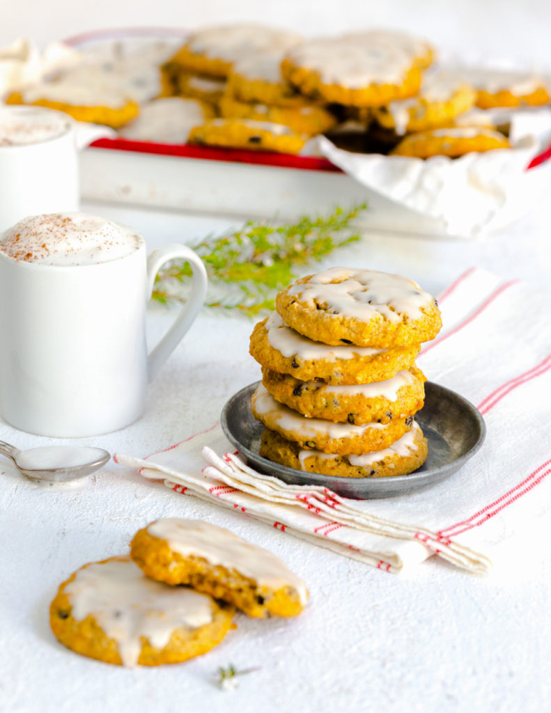 A stack of Iced Orange Currant Oatmeal Cookies with mugs filled with foam-topped coffee shot at an angle